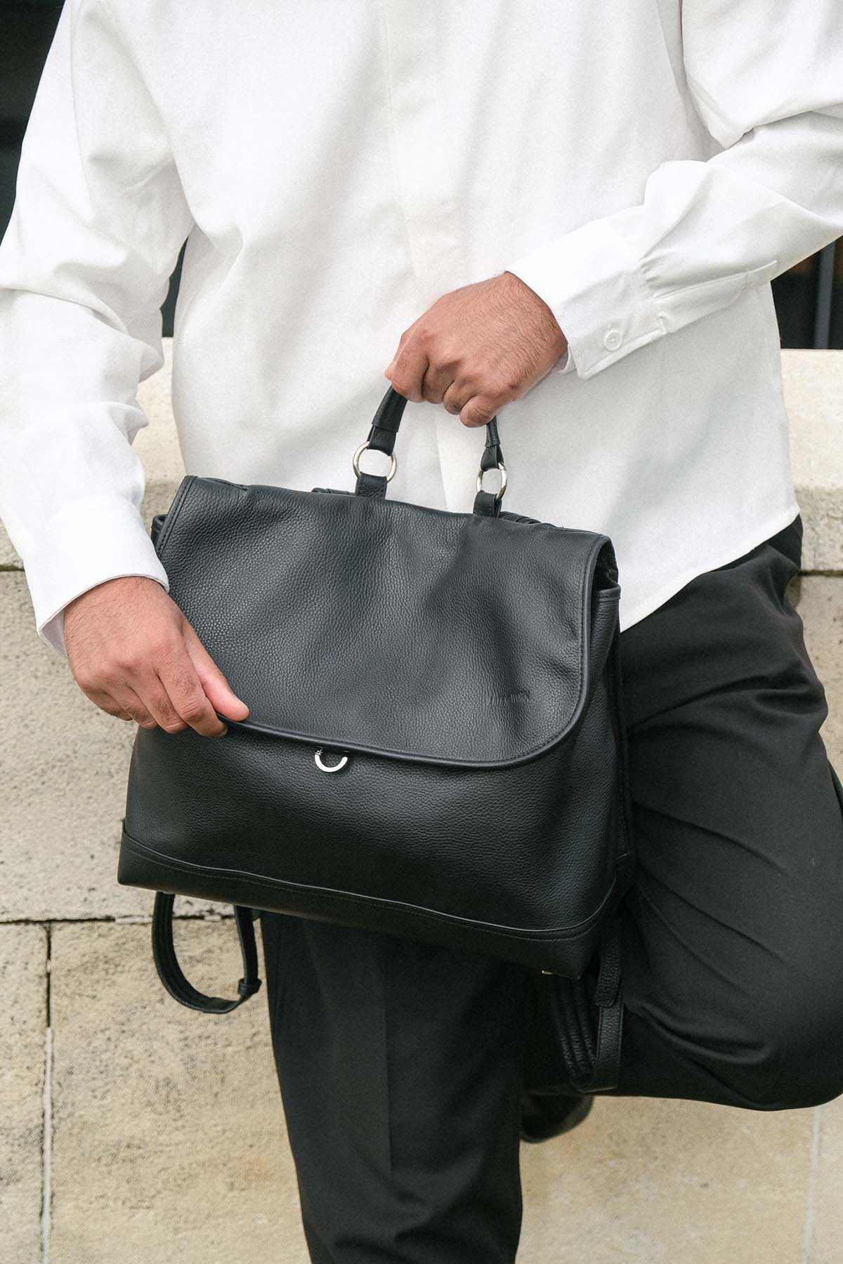 A person in a white shirt and black trousers holds a black leather backpack bag with top handles and a small circular metal ring on the front flap.