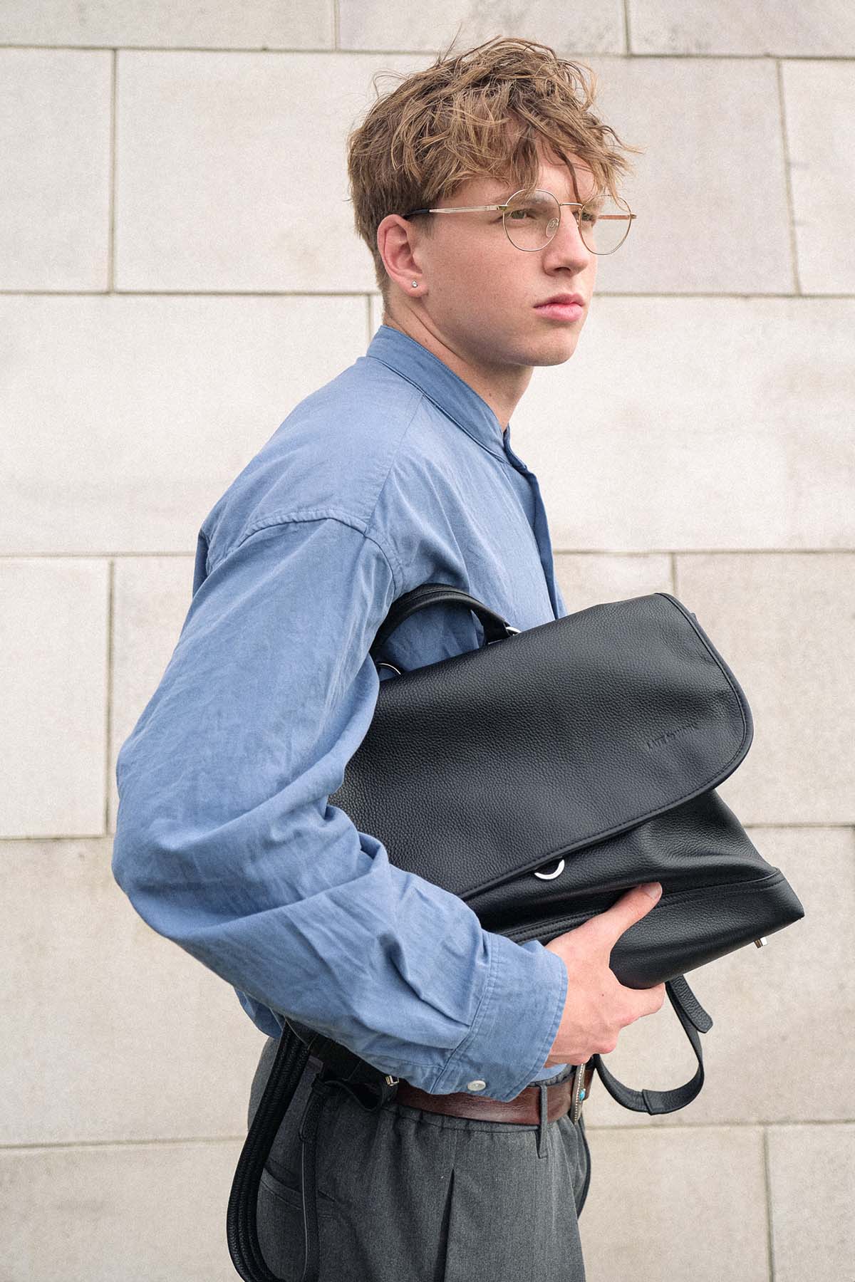 A man in a blue shirt and glasses carries a black leather backpack against his side, standing against a light stone wall.