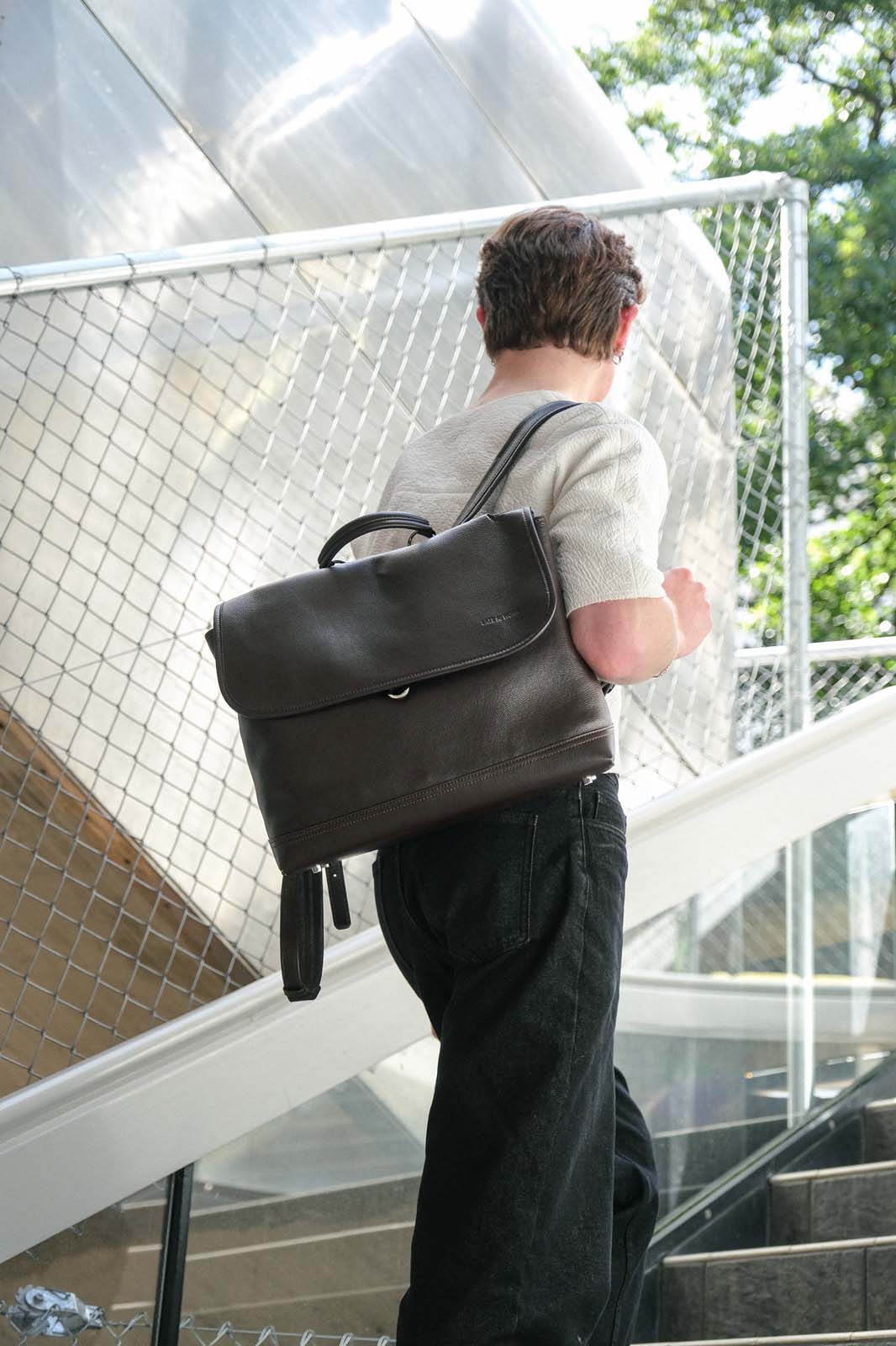 A man viewed from behind carrying a dark brown leather backpack over his shoulder while walking up outdoor stairs.