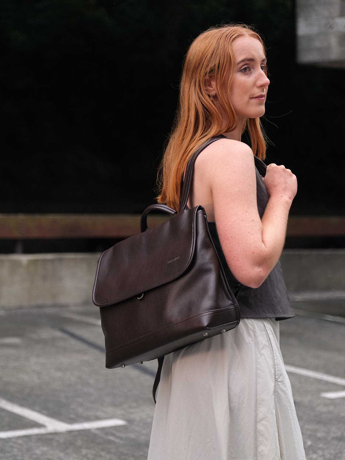 A woman with red hair wearing a dark gray sleeveless top and a light gray skirt carries a dark brown leather backpack over her shoulder in an outdoor parking lot setting.