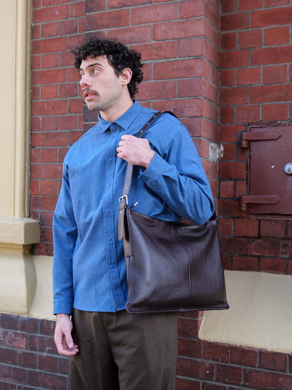 A man in a blue shirt wearing the brown Luxe Kite Bag on one shoulder while standing against a brick wall