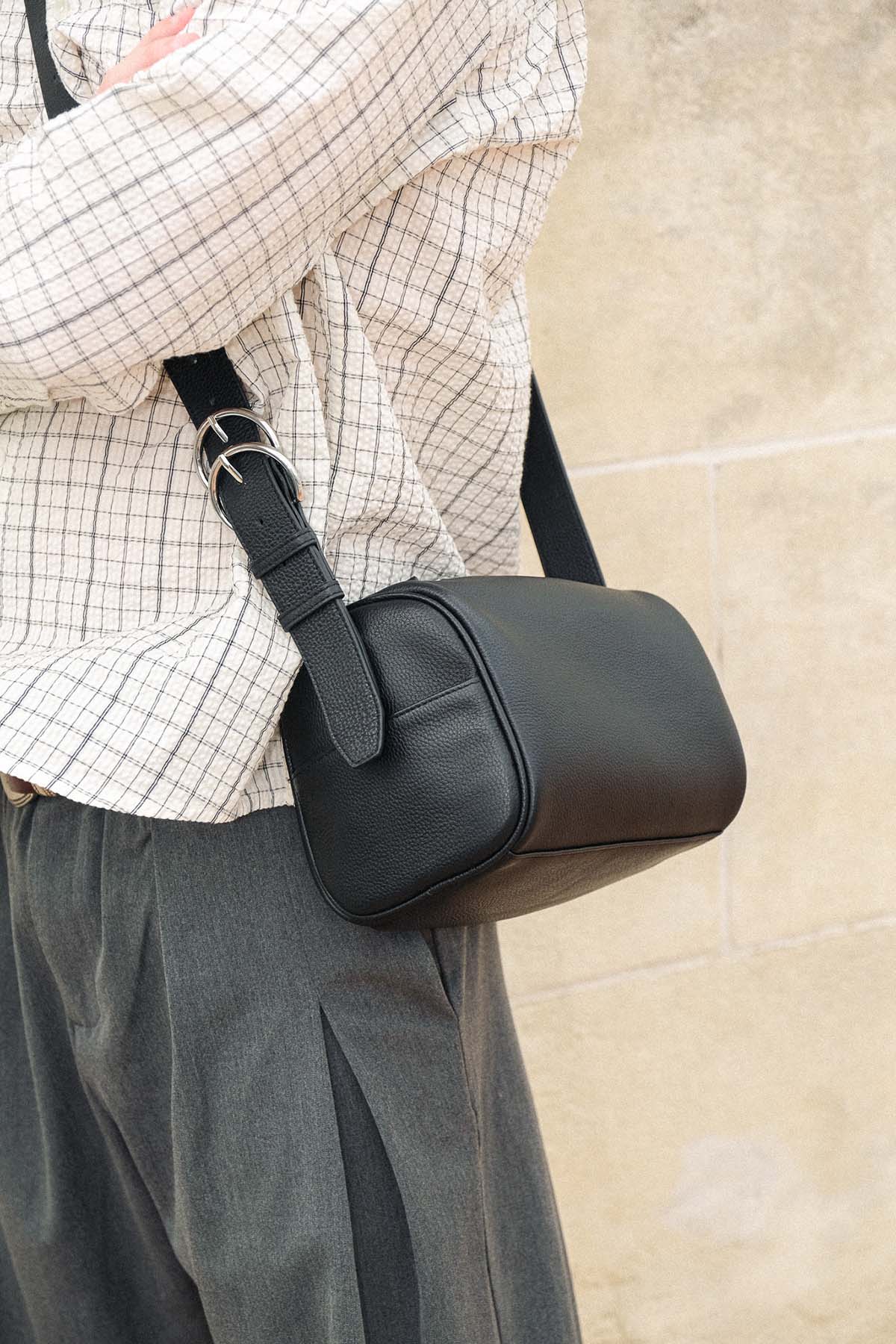 A person wearing a beige and black checkered shirt and dark gray trousers carries a small black leather crossbody bag with a wide strap and silver buckle against a beige stone wall.