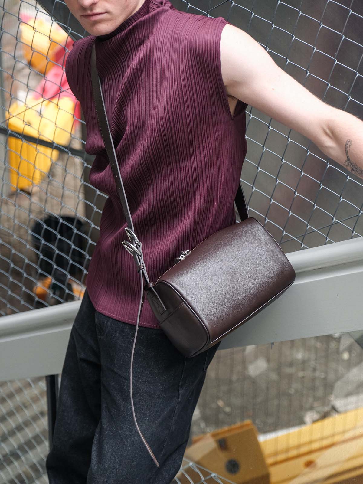 A person wearing a sleeveless ribbed burgundy top and dark jeans carries a dark brown leather crossbody bag with silver hardware, standing near a metal mesh fence.