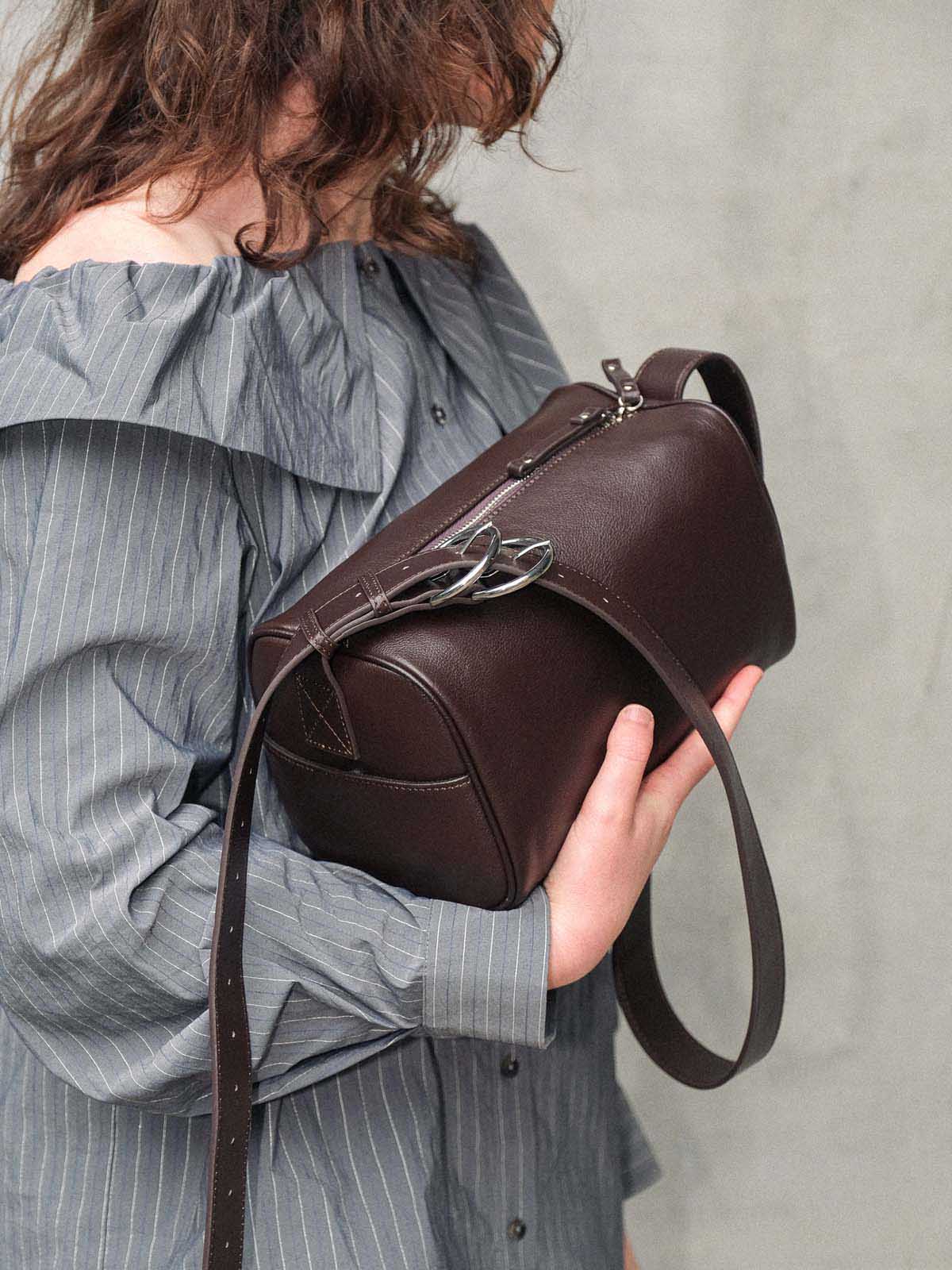 A person in a gray pinstripe off-the-shoulder blouse holds a dark brown leather shoulder bag with a silver-tone double buckle detail and a long strap against a textured gray wall.