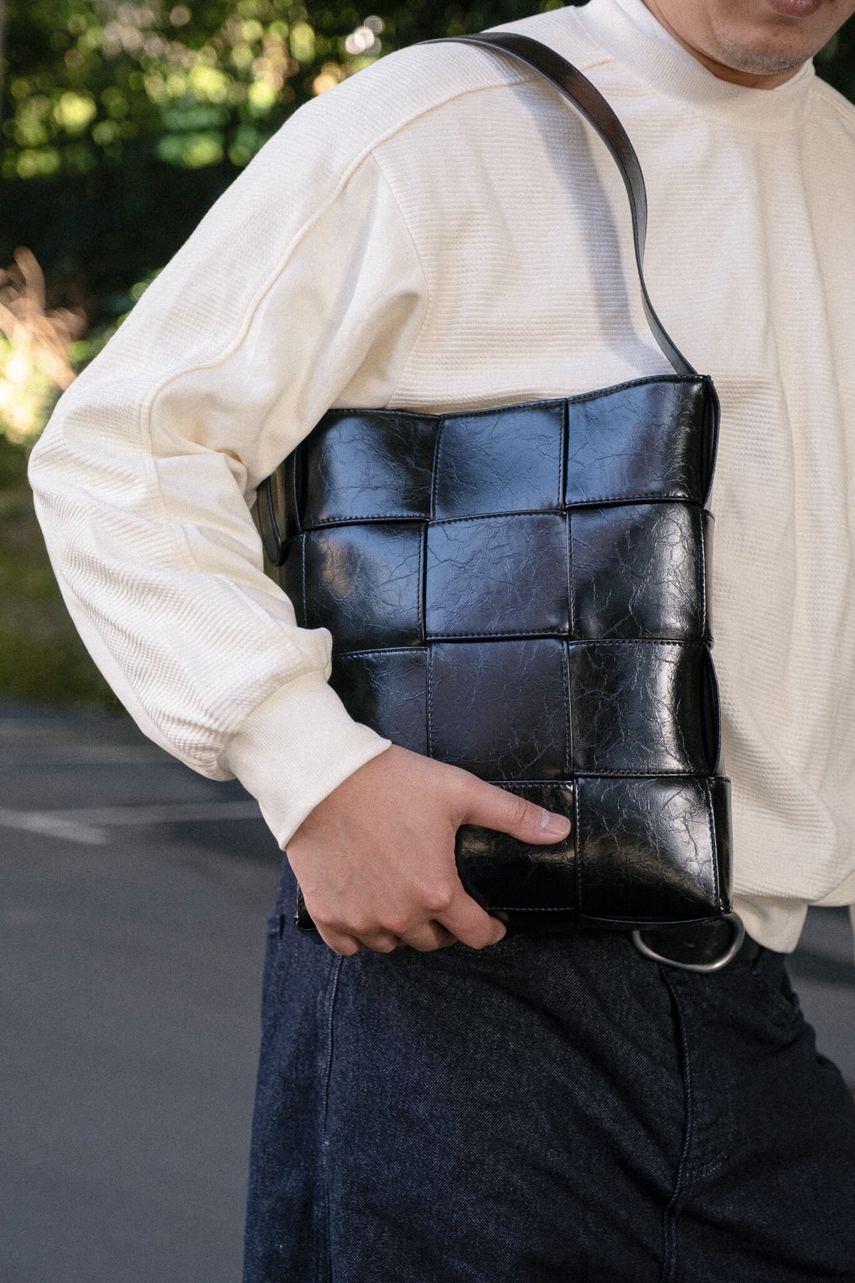 A person wearing a cream ribbed long-sleeve shirt and dark jeans carries a black woven leather shoulder bag outdoors.