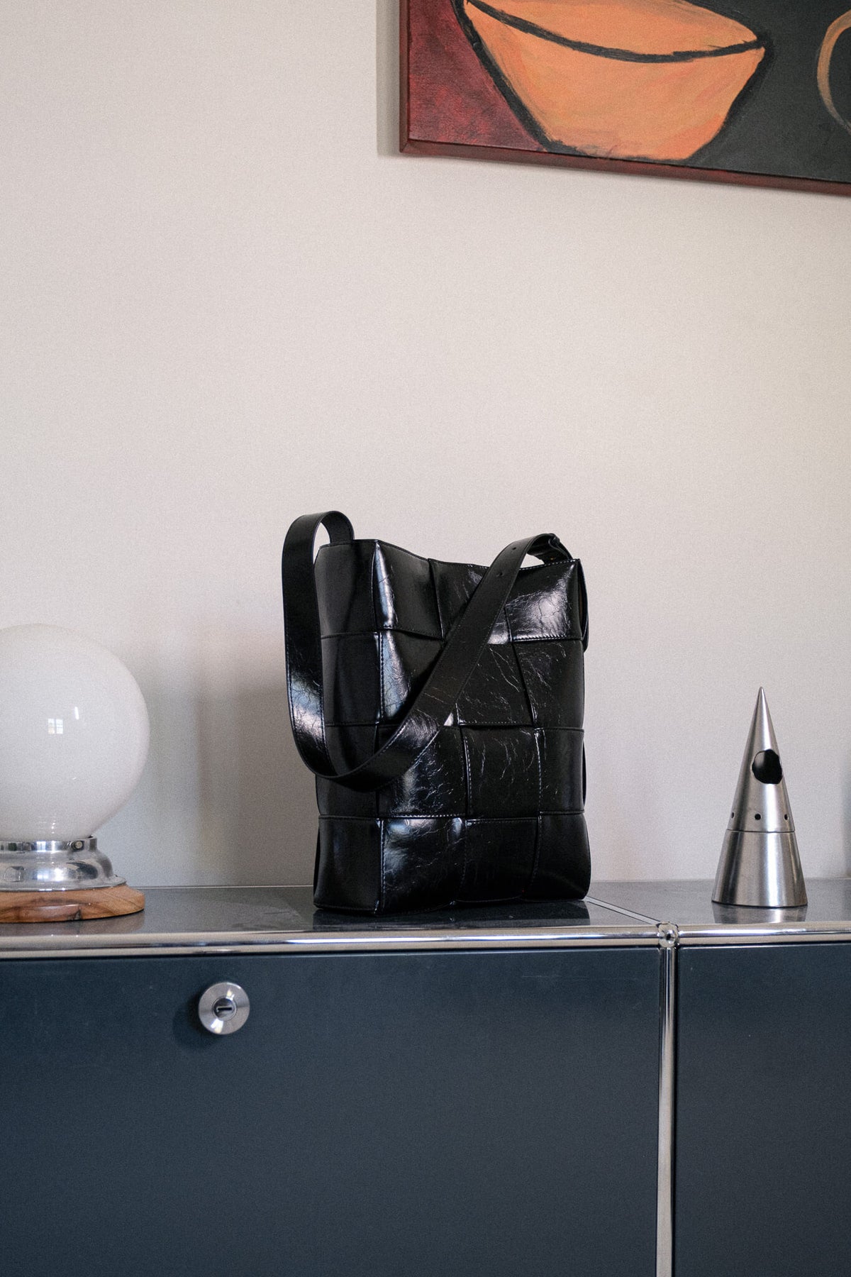 A black woven leather tote bag with a glossy finish, displayed on a dark blue-gray sideboard against a beige wall.
