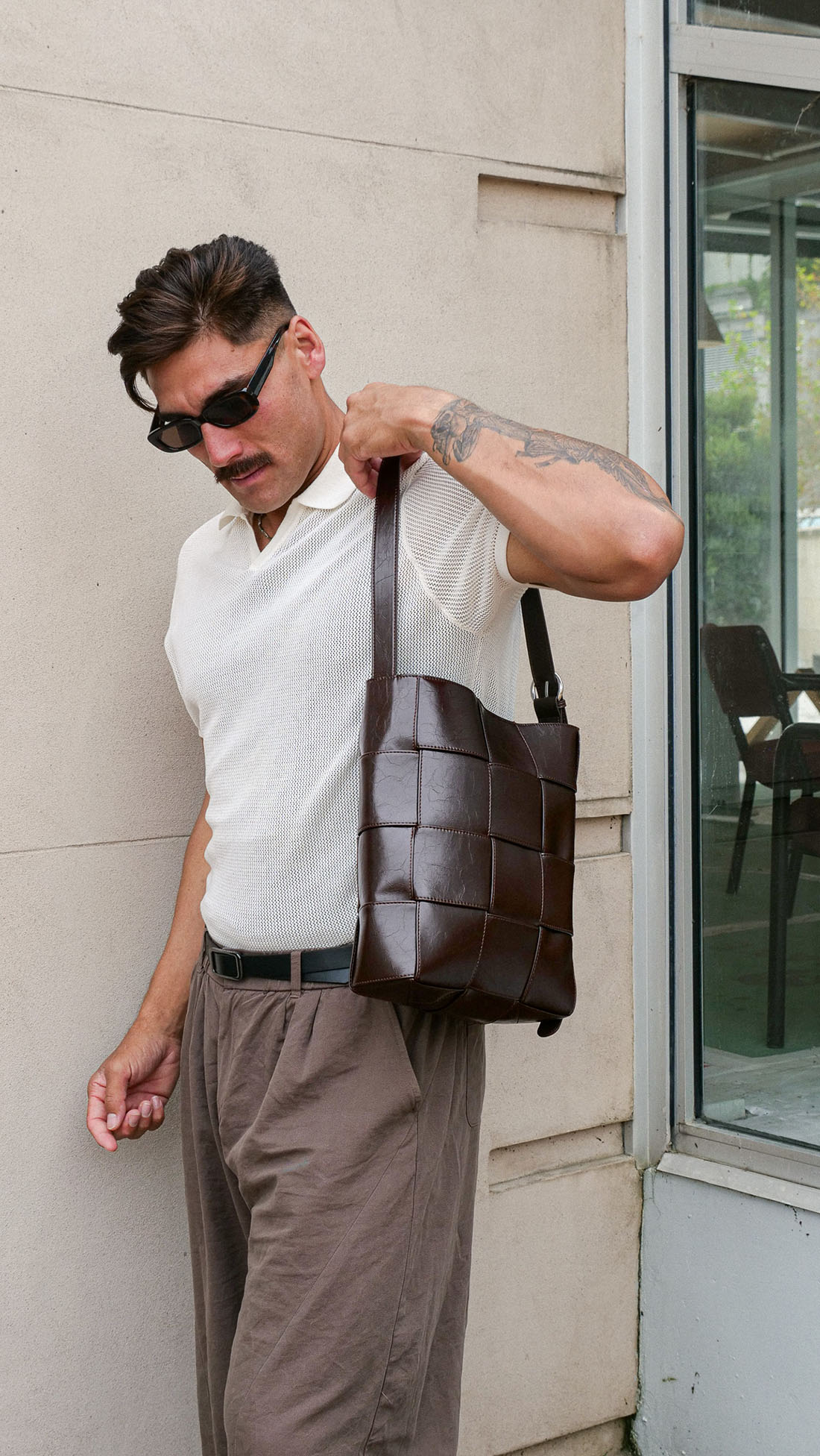 A man wearing a white polo shirt and brown trousers carries a dark brown woven leather shoulder bag against a beige wall.