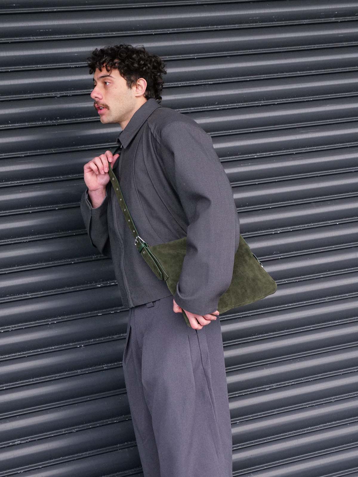 A man wearing a dark gray jacket and matching trousers stands in front of a dark gray metal shutter, carrying the medium Kite Bag in forest suede.