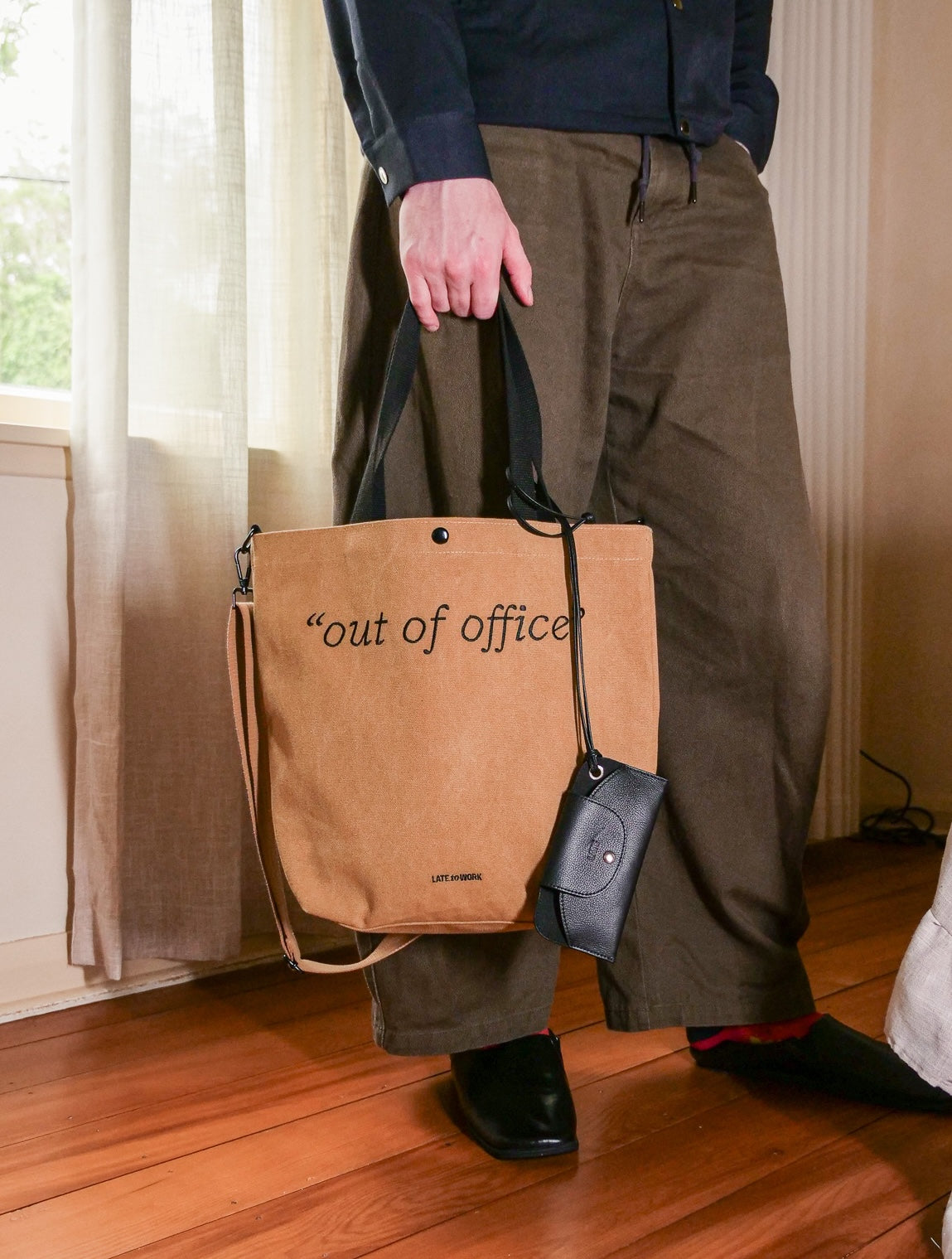 Person holding a brown tote bag with 'out of office' text, standing indoors.