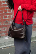 Person wearing a red sweater and holding a brown leather handbag against a brick wall.