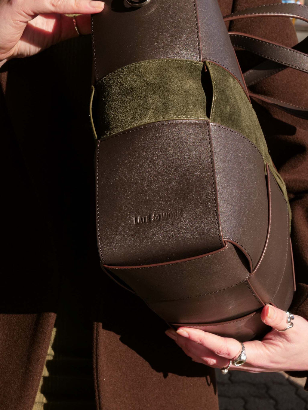 A close-up of a brown leather and suede shoulder bag held by hands outdoors. The bag features a dark brown leather body with an olive green suede accent panel and a debossed 'Late to Work' logo on the bag.