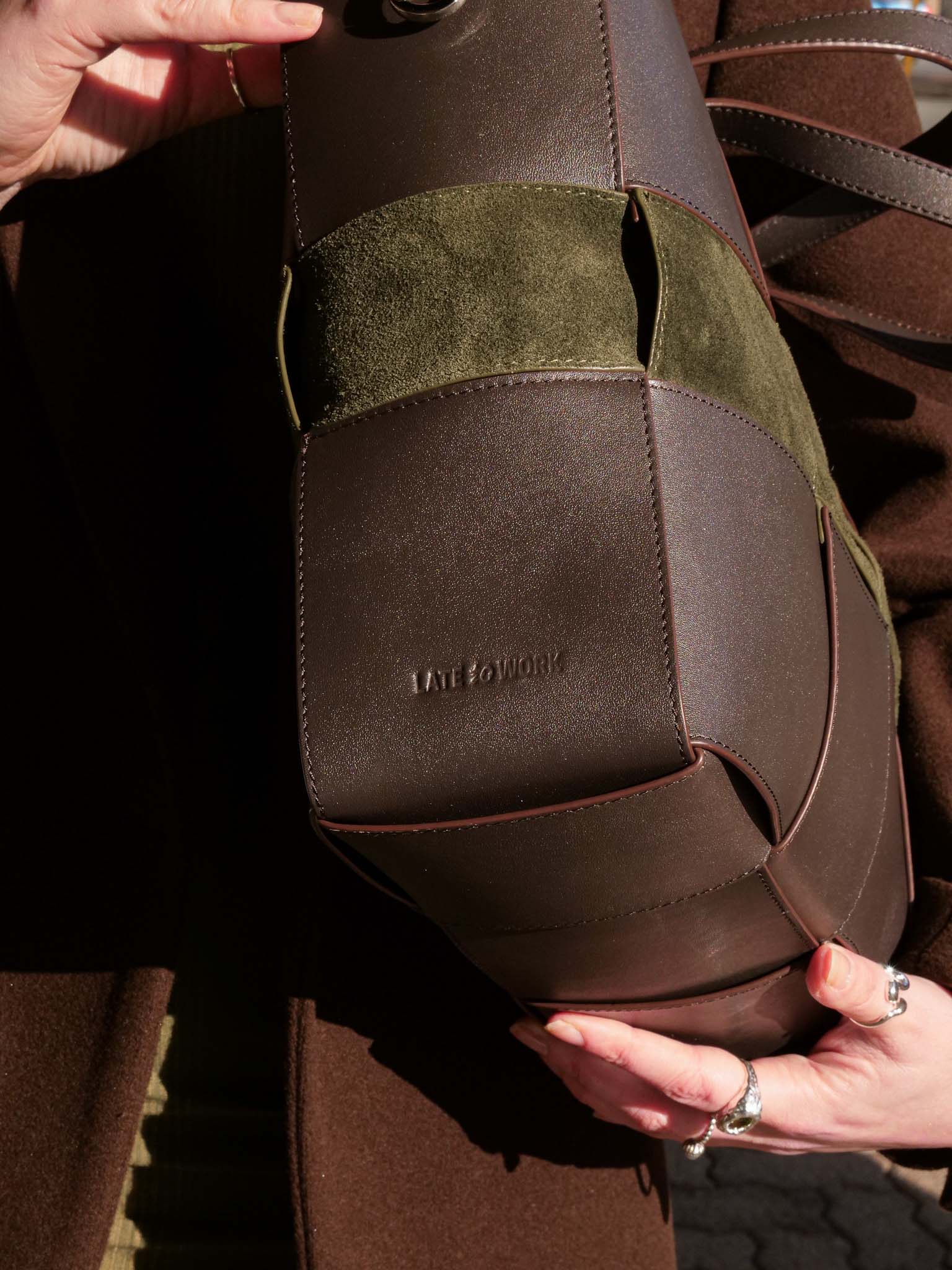 A close-up of a brown leather and suede shoulder bag held by hands outdoors. The bag features a dark brown leather body with an olive green suede accent panel and a debossed 'Late to Work' logo on the bag.