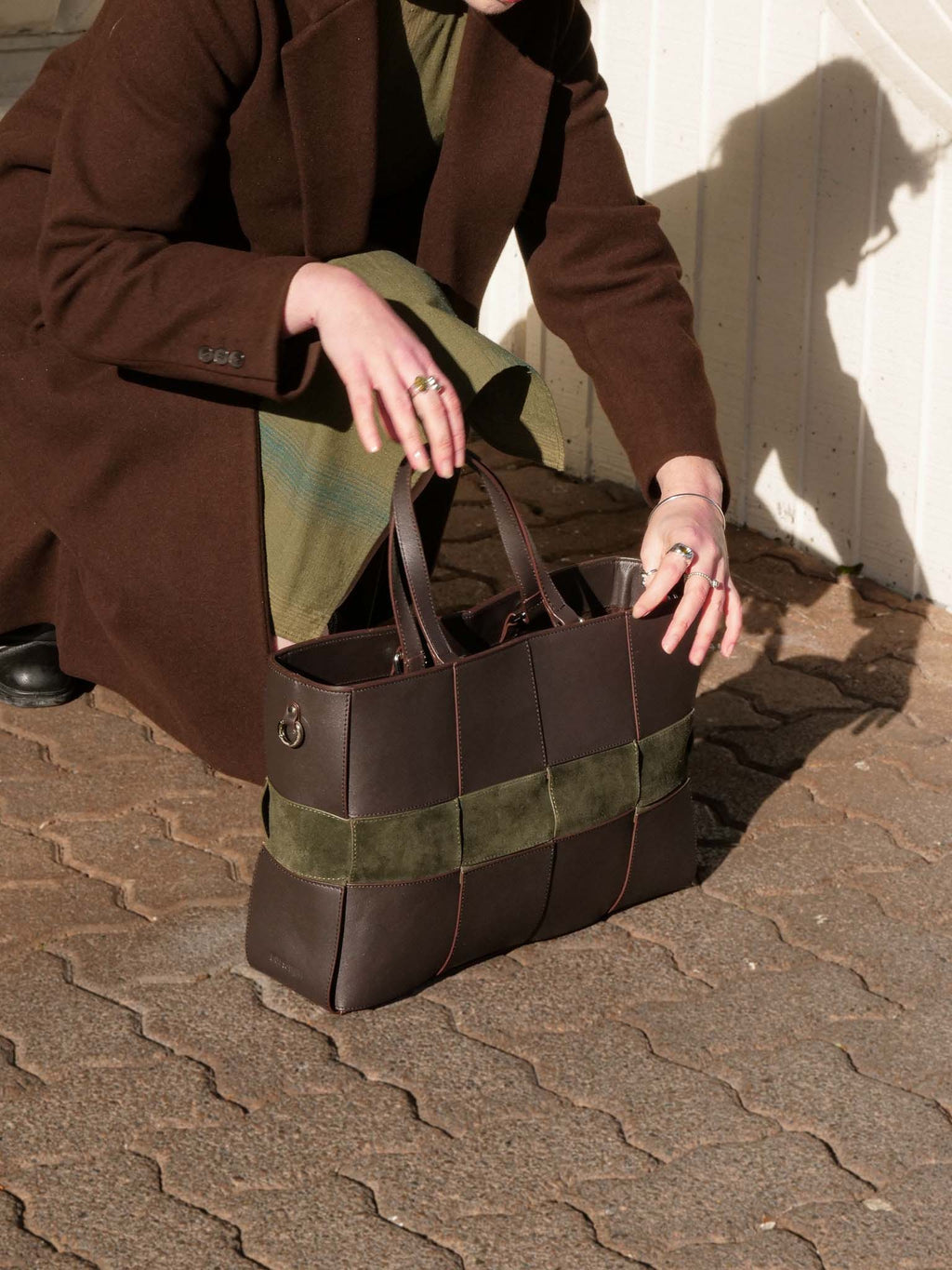 A person in a brown coat kneels outdoors on a paved surface, holding a structured brown leather tote bag with olive green suede-like horizontal panels.