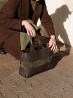 A person in a brown coat kneels outdoors on a paved surface, holding a structured brown leather tote bag with olive green suede-like horizontal panels.