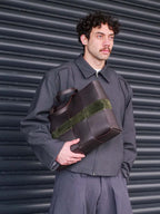 Man carrying the Reed Woven Bag in Forest and Walnut under one arm against a dark wall