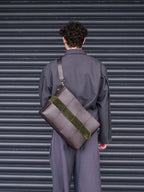 A person viewed from behind wearing a dark gray suit and carrying a large structured brown leather shoulder bag with a wide olive green suede-like band across the front, standing in front of a dark gray corrugated metal shutter.