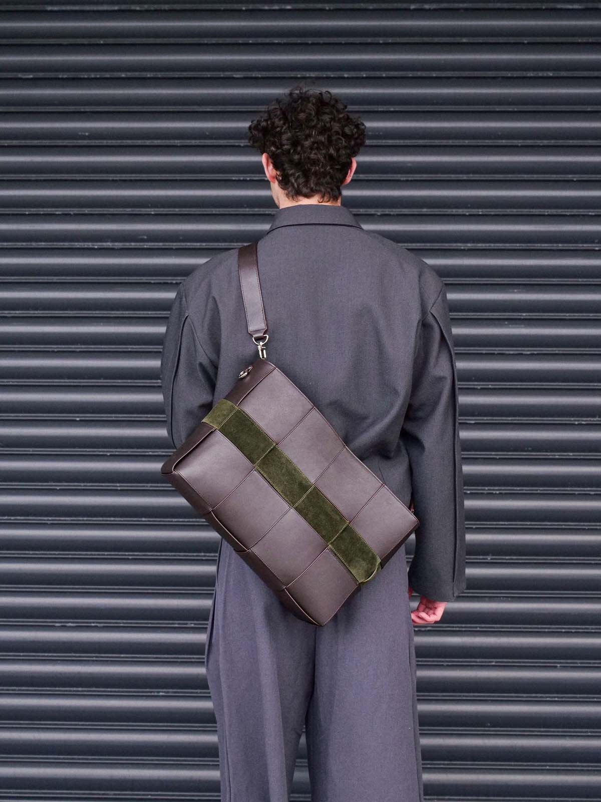 A person viewed from behind wearing a dark gray suit and carrying a large structured brown leather shoulder bag with a wide olive green suede-like band across the front, standing in front of a dark gray corrugated metal shutter.