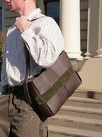 A man wearing a white pinstripe shirt and brown pants carries a brown leather messenger bag with an olive green suede accent strap, standing on stone steps in front of a building with white columns.