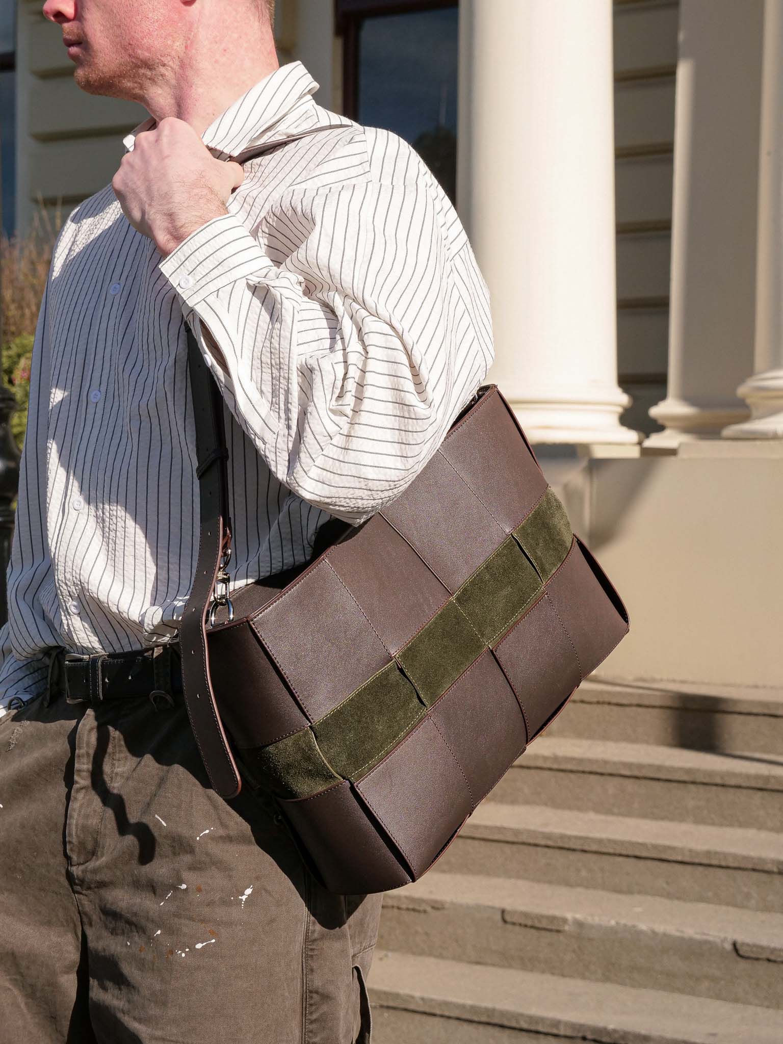 A man wearing a white pinstripe shirt and brown pants carries a brown leather messenger bag with an olive green suede accent strap, standing on stone steps in front of a building with white columns.