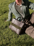 A person sitting on grass outdoors beside a structured brown leather tote bag with a wide olive green band and dark handles. The person wears a green cardigan, striped shirt, and brown pants.