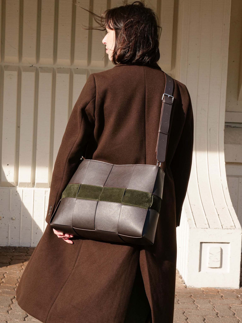 A woman wearing a brown coat is shown from behind, carrying a large structured shoulder bag with a gray leather body, a green woven accent band, and a dark strap, set against a cream-colored wooden building background.