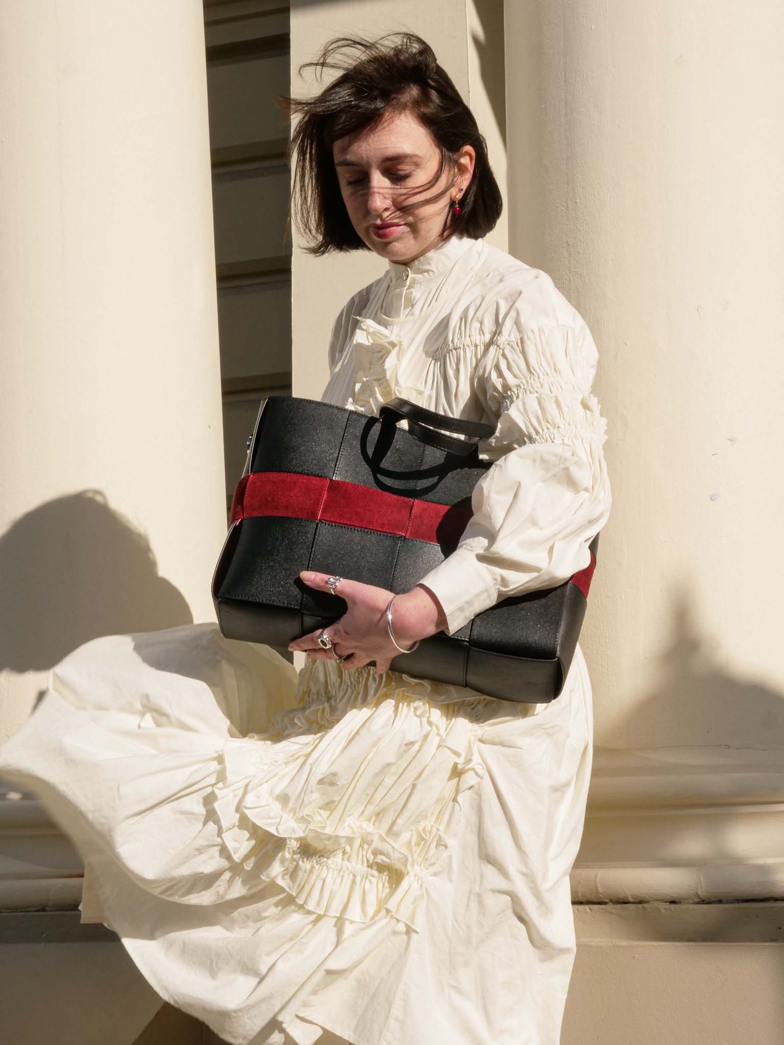 A woman in a cream ruffled dress holds a black leather tote bag with a bold red stripe, posed against a warm beige stone wall.