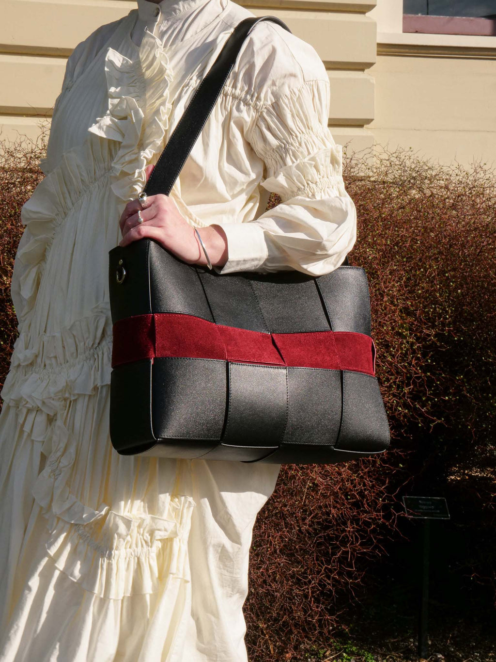 A person in a cream ruffled dress carries a structured black handbag with a wide red horizontal band and a black shoulder strap, photographed outdoors against a beige building and brown shrubs.