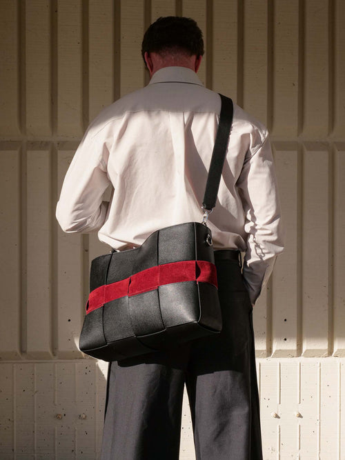 A man viewed from behind wearing a white shirt and dark trousers, carrying a black leather shoulder bag with a bold red horizontal stripe against a beige corrugated metal wall.