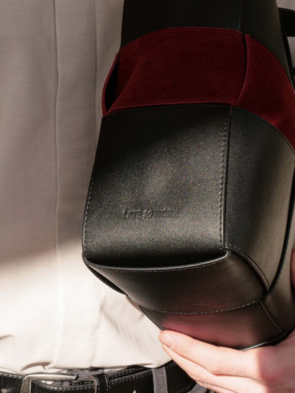 A close-up of a hand holding a structured black leather bag with a deep burgundy velvet panel near the top, against a light beige shirt background.