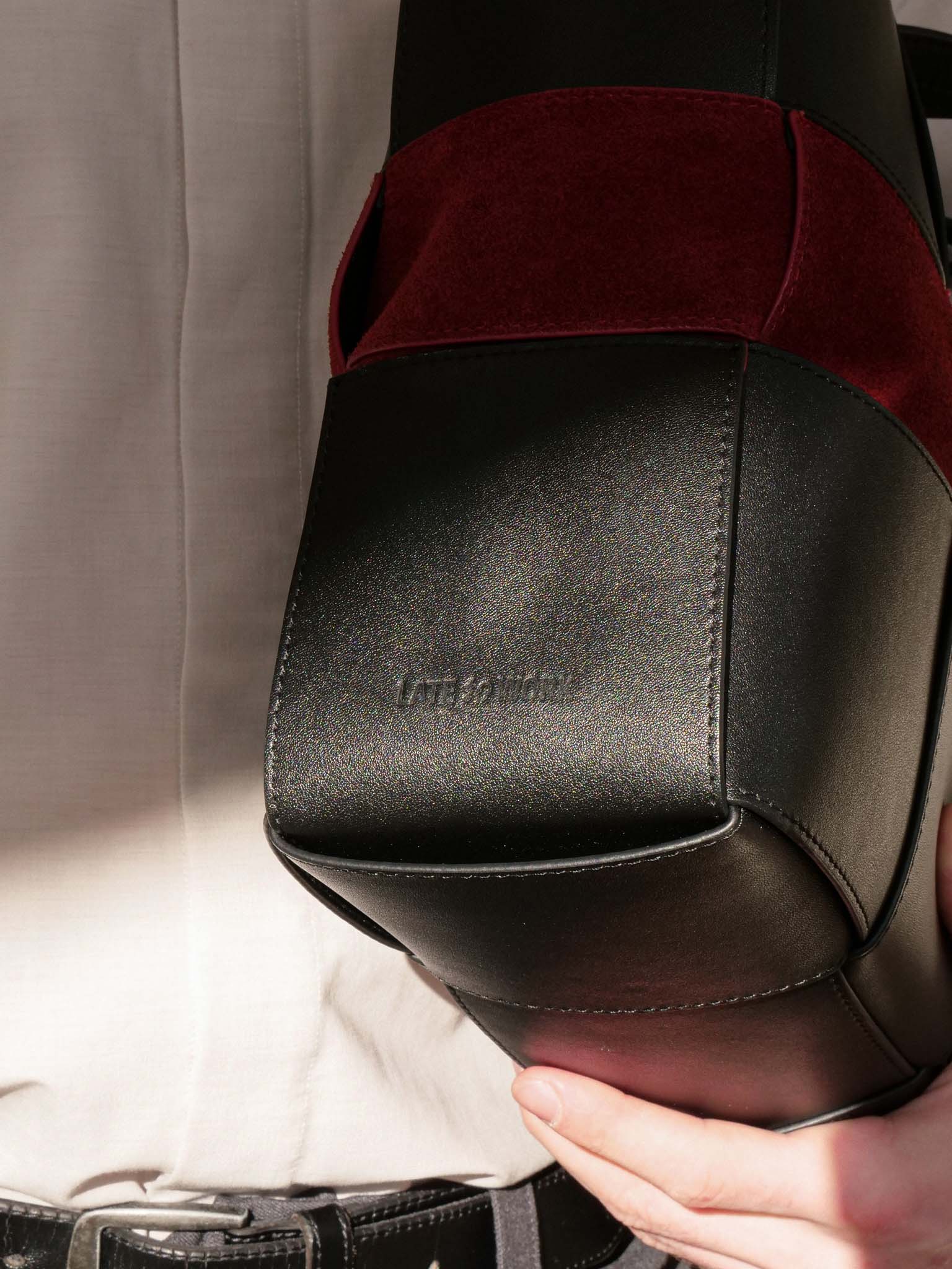 A close-up of a hand holding a structured black leather bag with a deep burgundy velvet panel near the top, against a light beige shirt background.