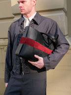 A man in a dark gray jacket and trousers carries a large black leather handbag with a bold red suede stripe across the front, set against a beige stone building background.