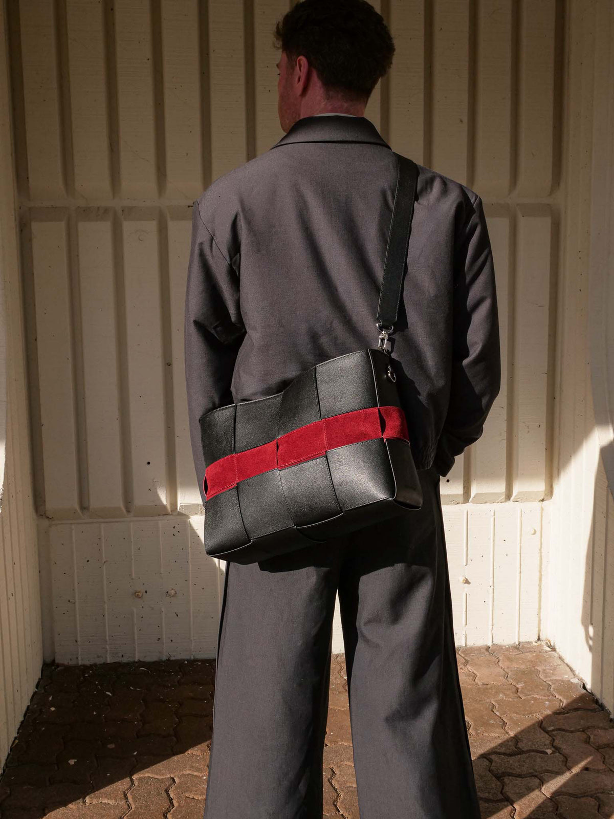 A man viewed from behind wearing a dark gray suit and carrying a black leather shoulder bag with a bold red horizontal stripe, standing against a cream-colored paneled wall.