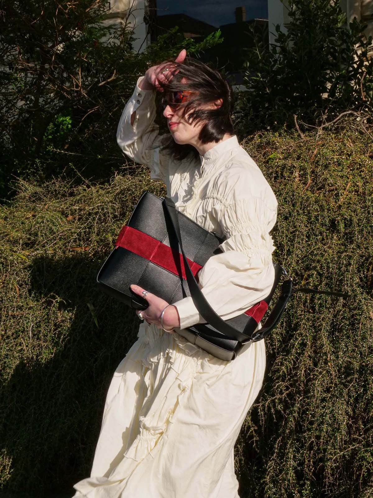 A person outdoors wearing a cream puff-sleeve dress and holding a black tote bag with a bold red horizontal stripe and black strap, set against a background of green foliage.