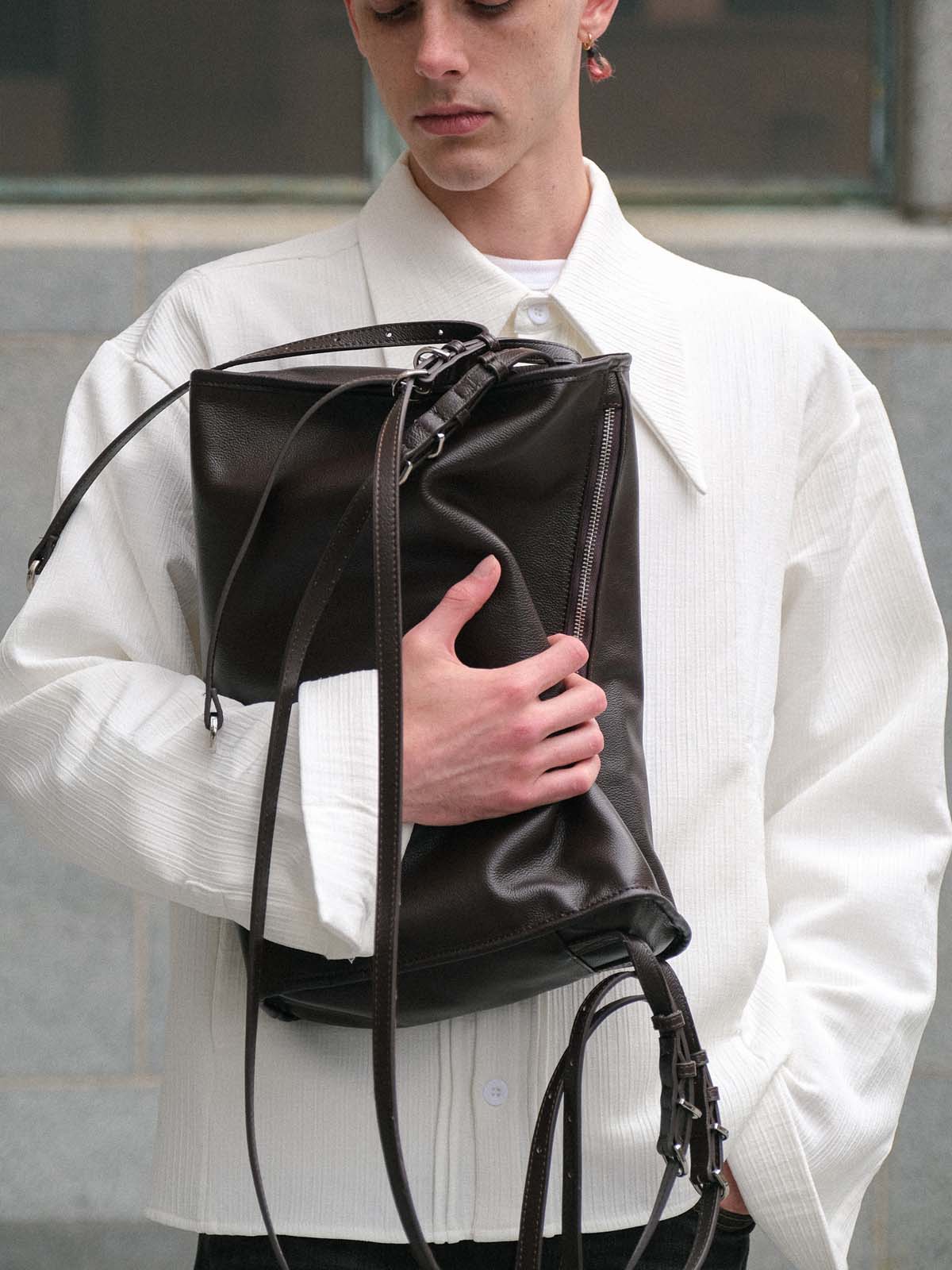 A model wearing a white textured shirt holds a large black leather bag with long straps.