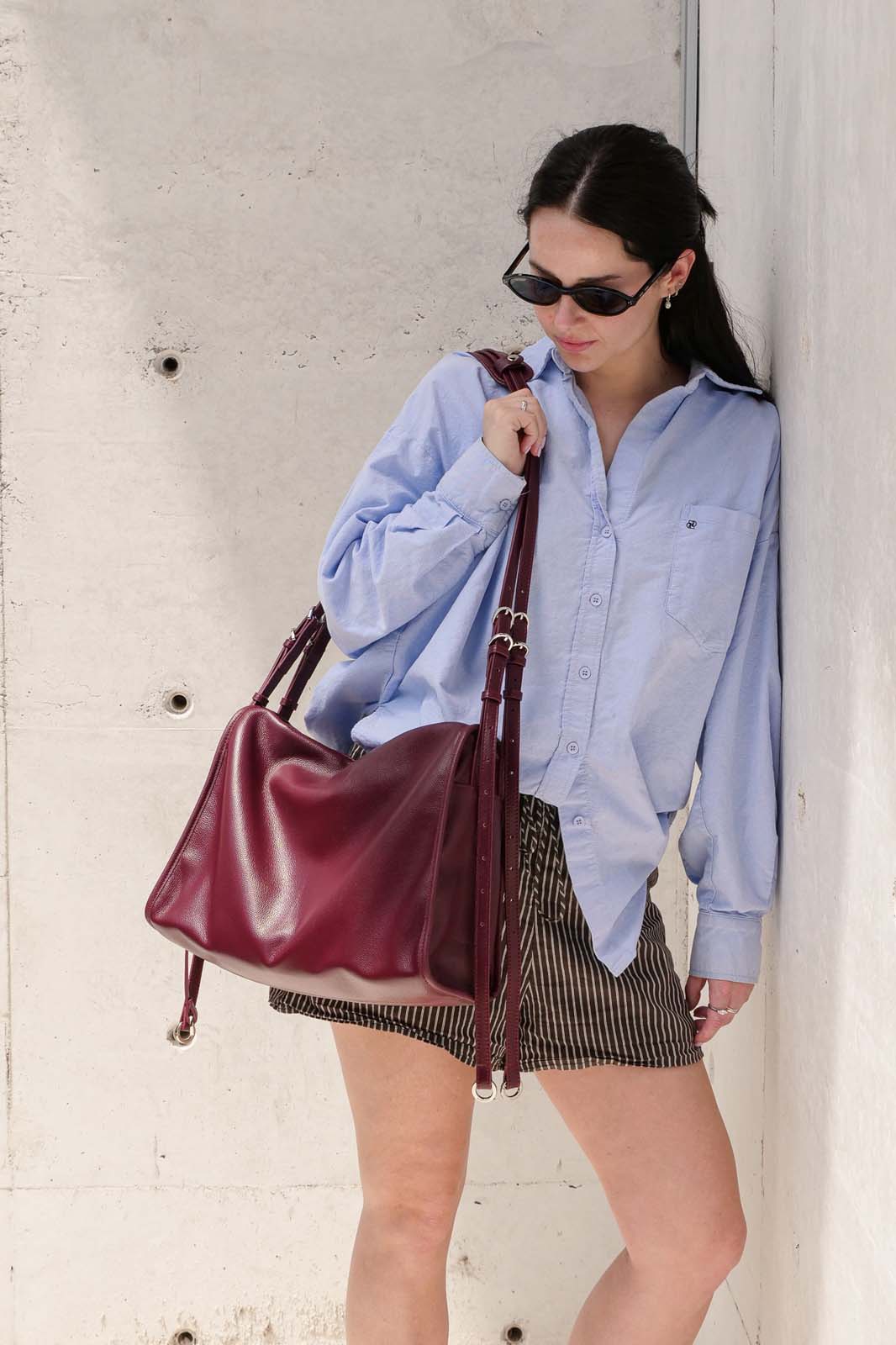 A woman wearing a light blue button-up shirt, black sunglasses, and a black-and-white striped mini skirt carries the burgundy Willow bag against a textured concrete wall.