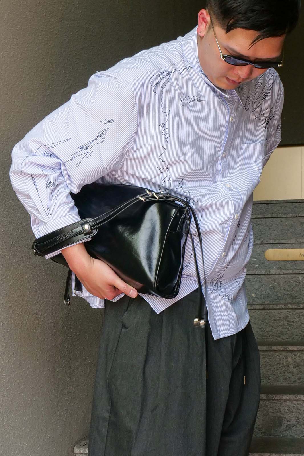 A man wearing a white long-sleeve button-up shirt with black line-art illustrations, dark gray wide-leg trousers, and sunglasses, holding a black leather shoulder bag.