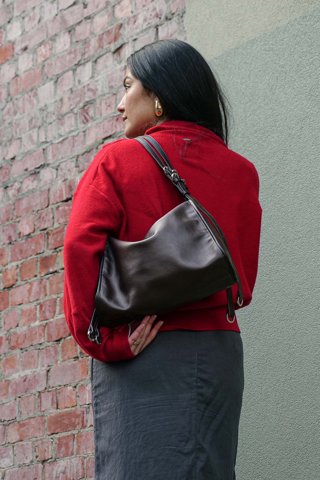 A woman wearing a red sweater and dark gray skirt carries a dark brown leather shoulder bag against her back, standing in front of a brick wall and a gray wall.