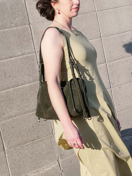 Woman carrying the small Willow Bag in forest suede on her shoulder while standing in front of a concrete wall