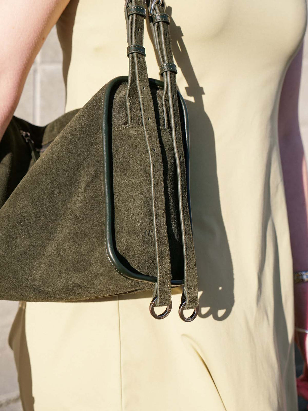 Close-up of a person wearing a cream-colored dress and carrying an olive green suede shoulder bag with dark trim and long straps.