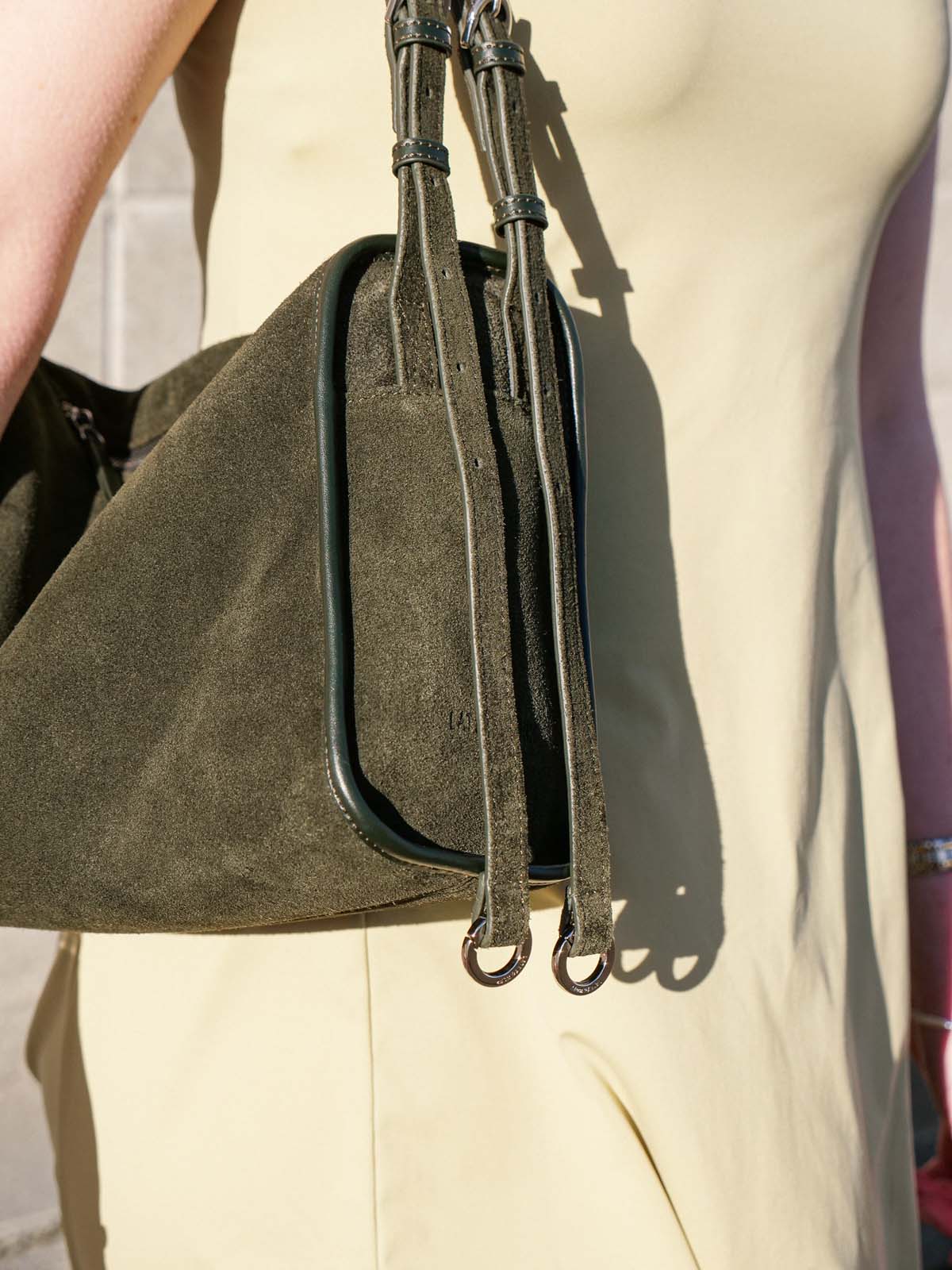 Close-up of a person wearing a cream-colored dress and carrying an olive green suede shoulder bag with dark trim and long straps.