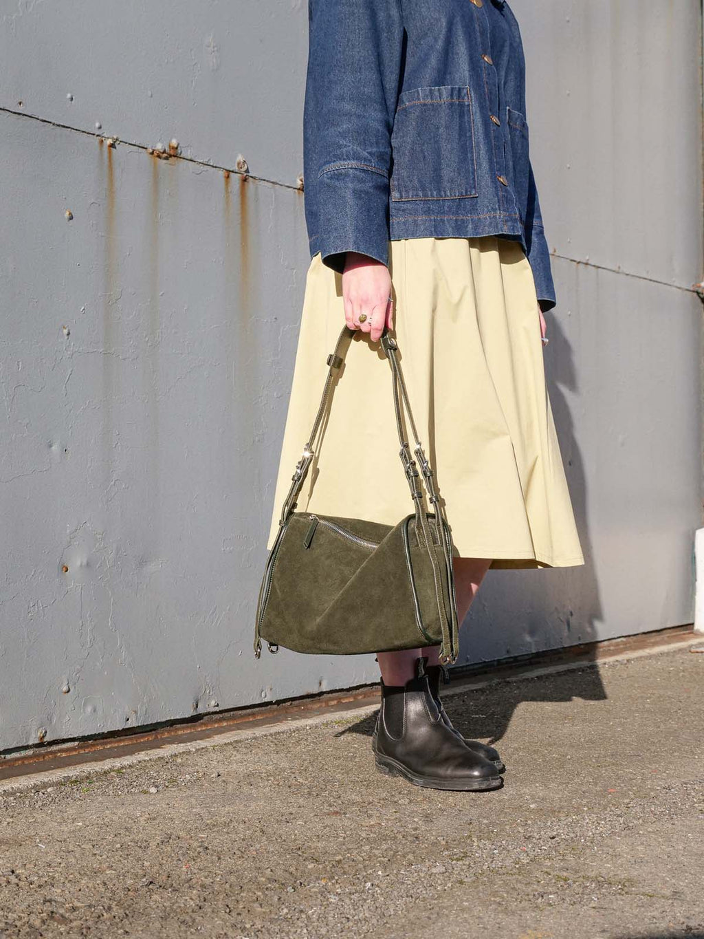 A person wearing a blue denim jacket, a light beige midi skirt, and dark brown Chelsea boots holds an olive green suede shoulder bag against a gray metal wall.