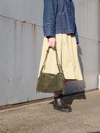 A person wearing a blue denim jacket, a light beige midi skirt, and dark brown Chelsea boots holds an olive green suede shoulder bag against a gray metal wall.