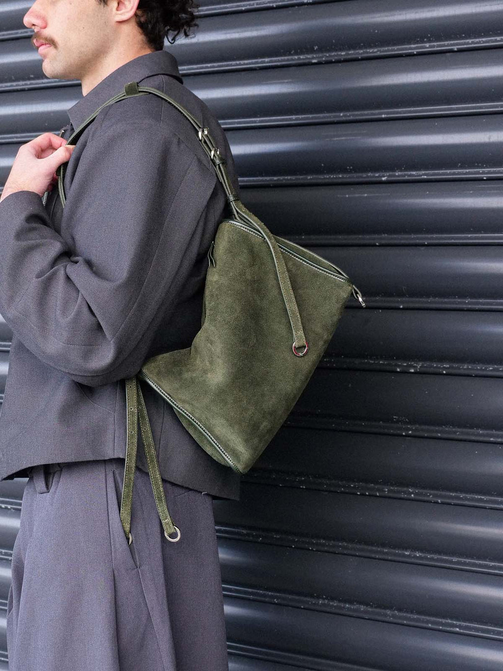 A person wearing a dark gray jacket and matching pants carries an olive green suede shoulder bag with a matching strap against a dark corrugated metal wall.
