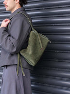 A person wearing a dark gray jacket and matching pants carries an olive green suede shoulder bag with a matching strap against a dark corrugated metal wall.
