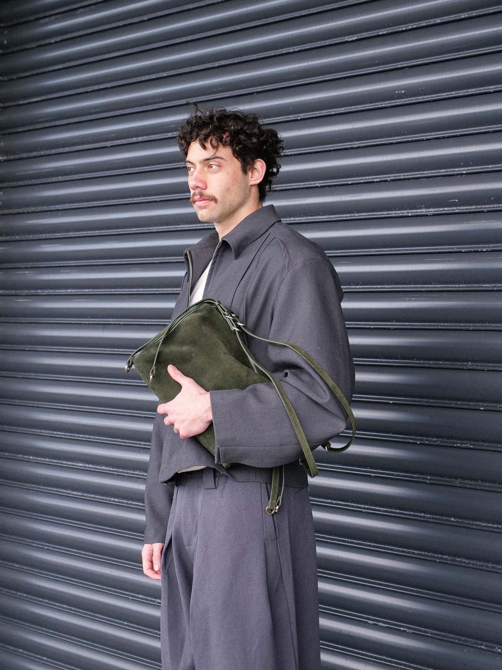 A man wearing a dark gray oversized jacket and matching trousers stands in front of a dark gray metal shutter, holding an olive green suede leather shoulder bag.