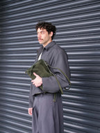 A man wearing a dark gray oversized jacket and matching trousers stands in front of a dark gray metal shutter, holding an olive green suede leather shoulder bag.