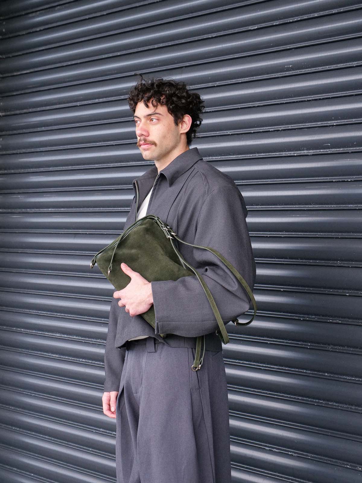 A man wearing a dark gray oversized jacket and matching trousers stands in front of a dark gray metal shutter, holding an olive green suede leather shoulder bag.