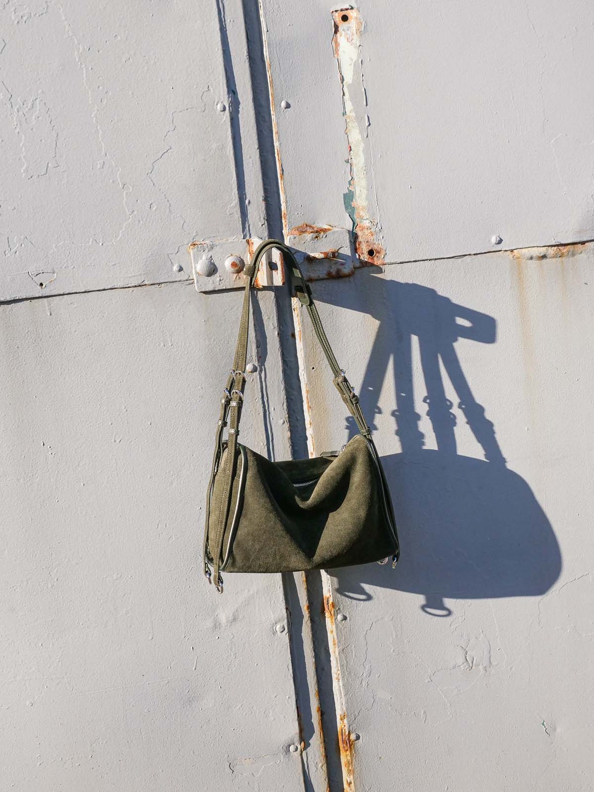 Small Willow Bag in forest suede hangs from a metal bracket on a weathered grey wall