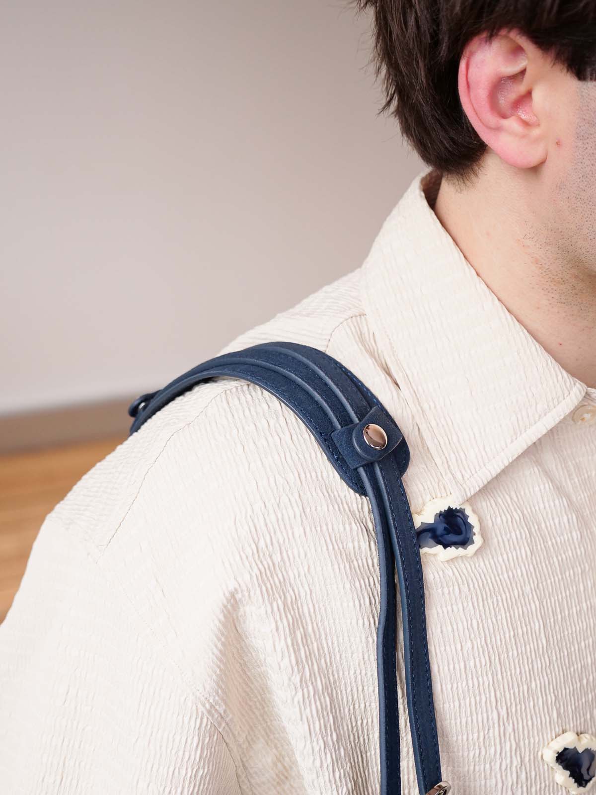 Close-up of a person wearing a cream textured shirt with dark blue suspenders over the shoulder, showing the strap detail and metal snap button.
